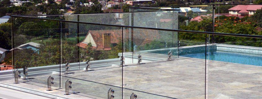 A rooftop swimming pool with glass fencing overlooking a neighborhood with trees and buildings.