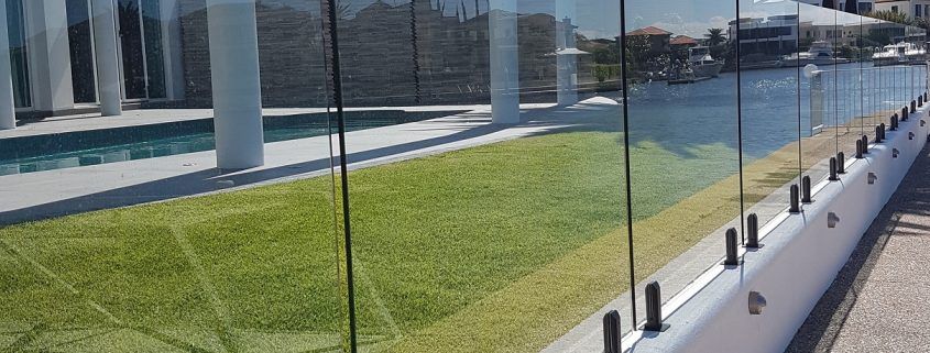 A glass fence with dark posts on a waterfront, separating a grassy area from the water.