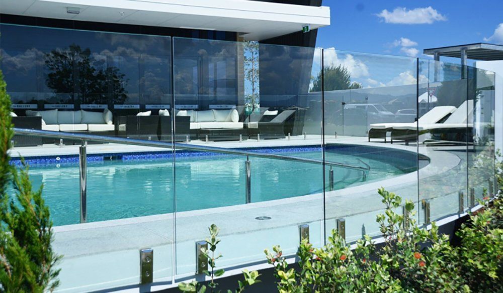 A pool surrounded by glass fencing; a building with outdoor seating on the other side, sunny day.