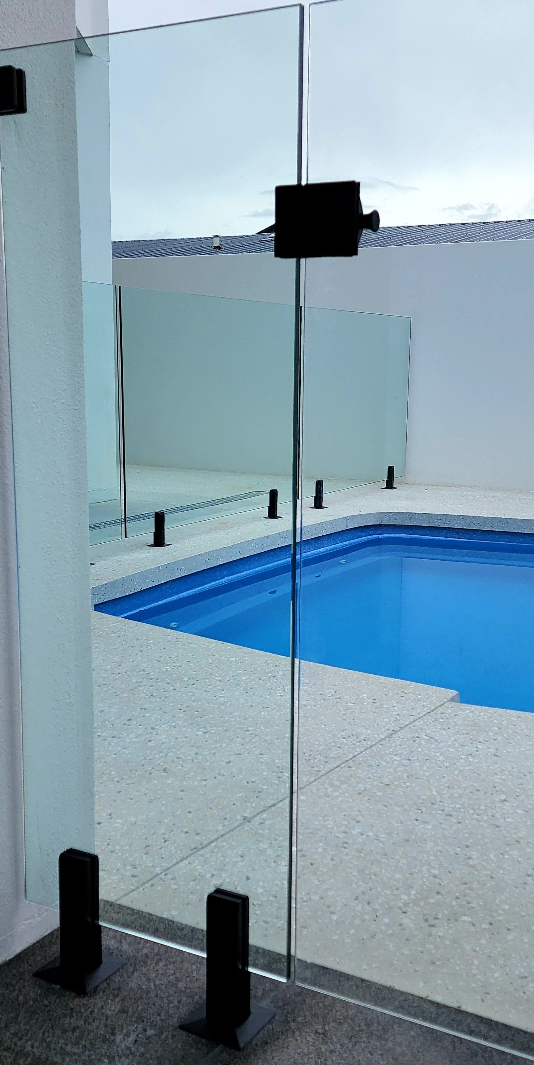 A glass pool fence with a black frame. The pool is visible through the transparent panels.