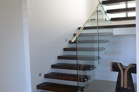 Dark wooden floating stairs with glass railing in a white-walled interior, gold handrail, with a dark-colored statue.