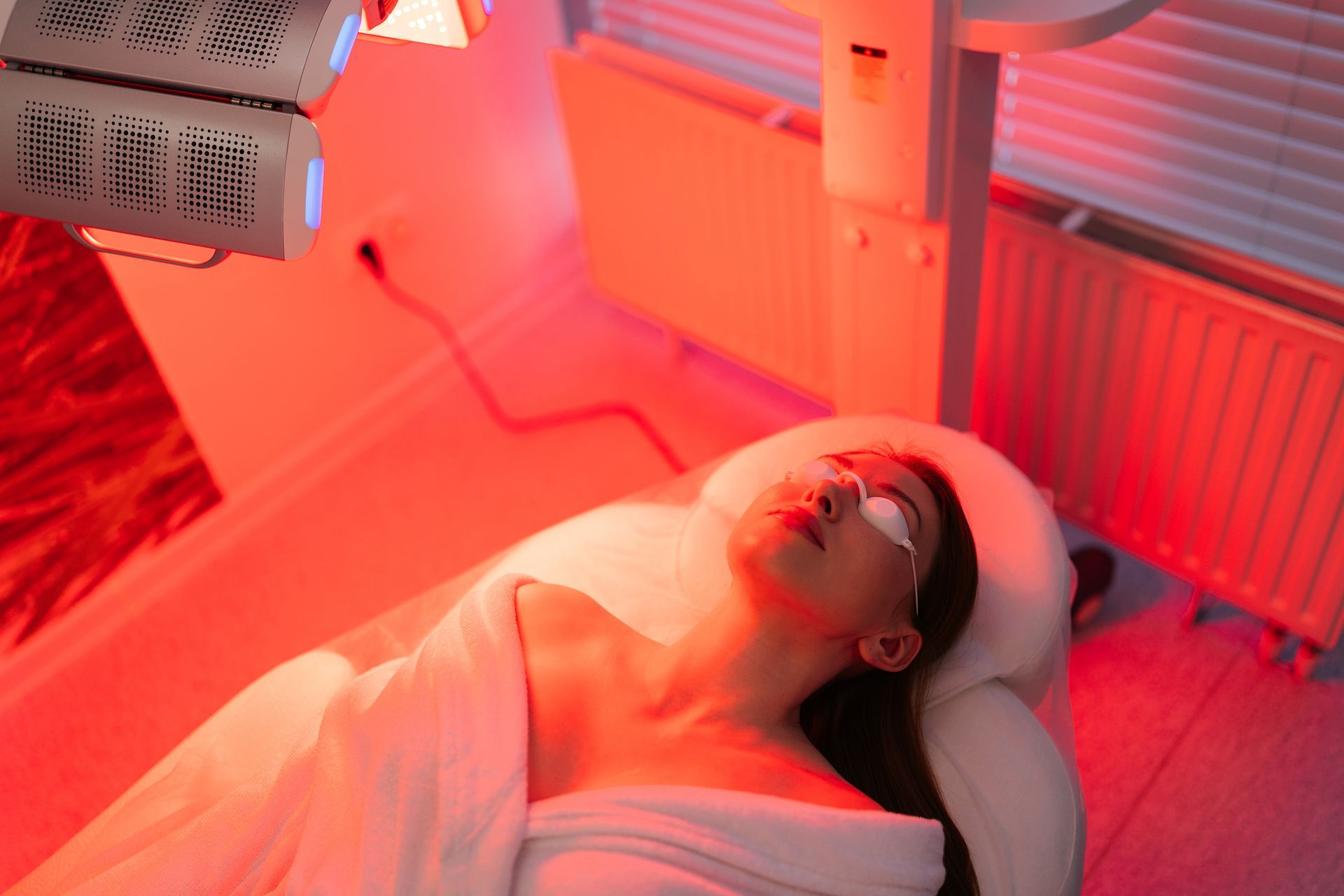 A woman is laying on a bed getting a red light treatment on her face.