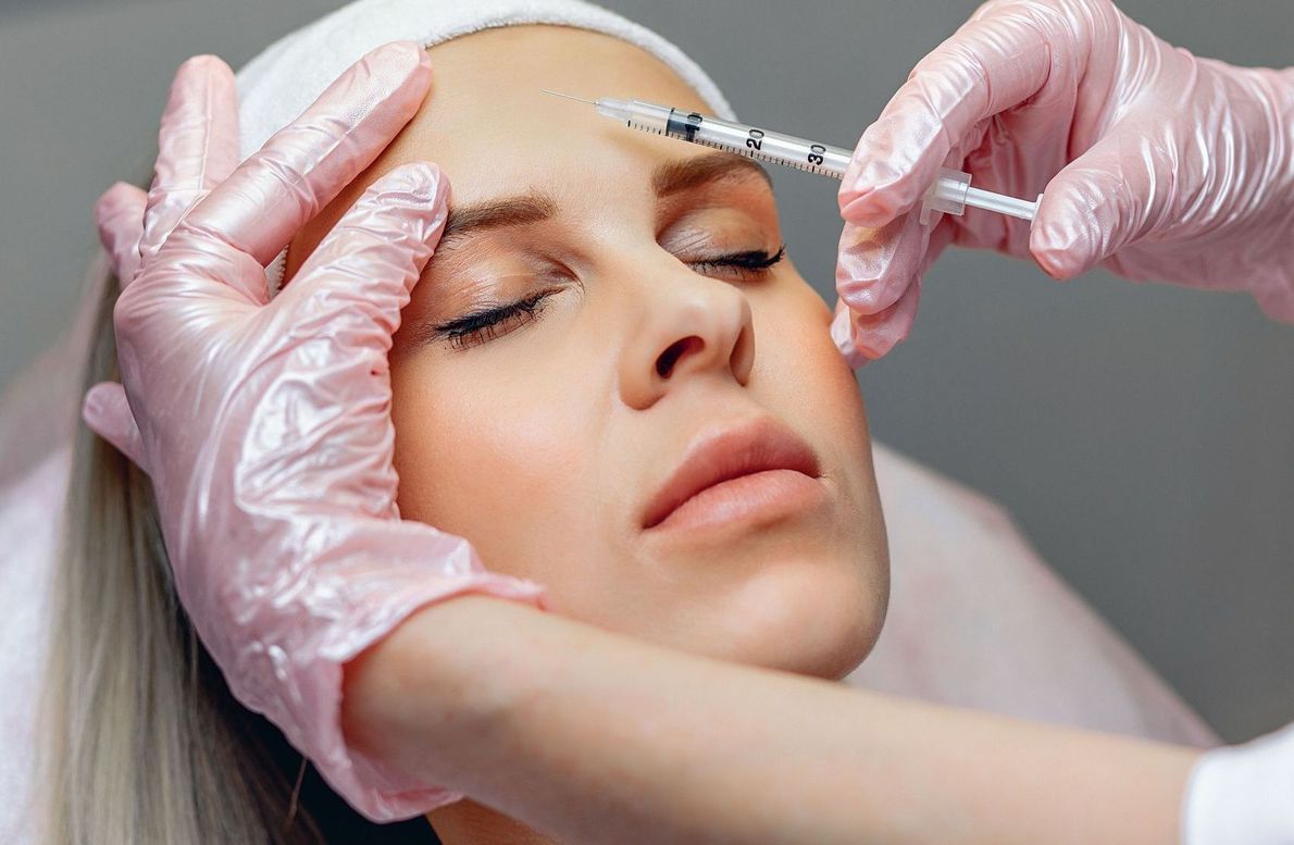 A woman receiving an injection in her forehead. Hands in pink gloves hold a syringe near her brow; she has her eyes closed.