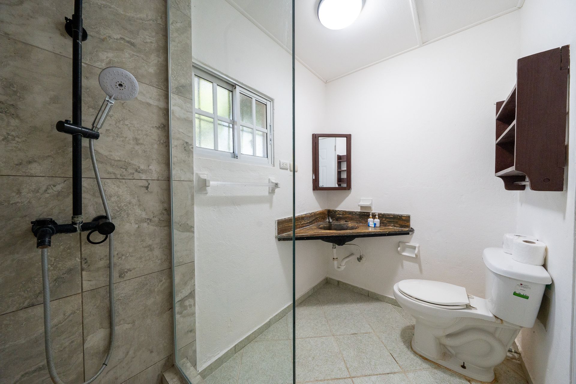 Small white bathroom with a shower, sink, toilet, mirror, and window.