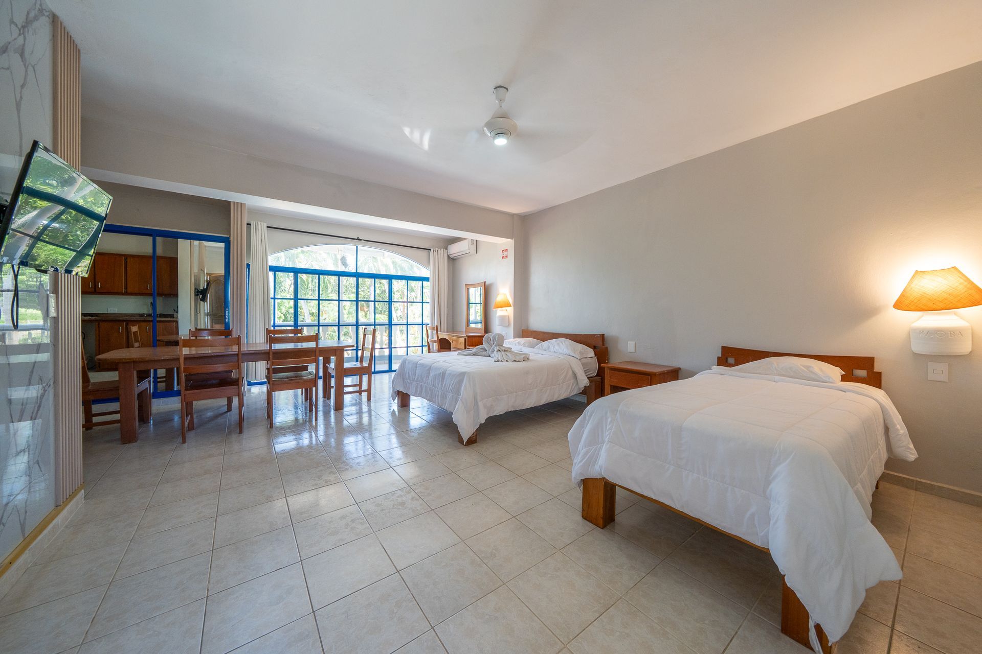 Bright hotel room with two beds, tiled floor, dining table, and sliding glass doors to a balcony