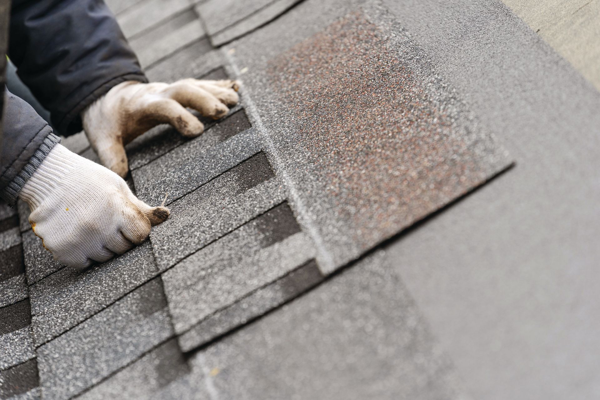 Close-up of a dark gray shingled roof with peeling edges beside a blurred suburban house and yard.