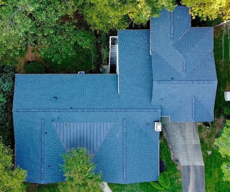 Aerial view of a large suburban house with gray shingle roof, front porch, and landscaped yard