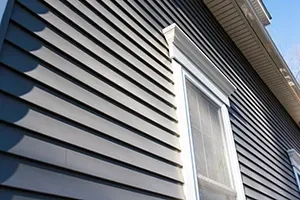 Close-up of gray house siding with a white-trimmed window and roof eaves against a blue sky