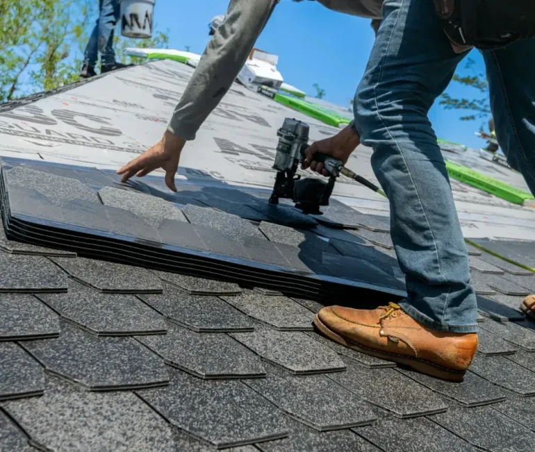 Worker installing gray shingles on a house roof with a nail gun.