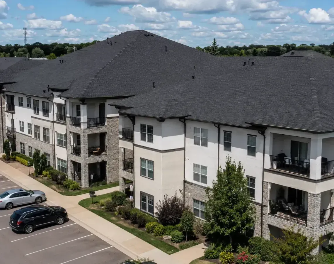 Aerial view of a white apartment complex with dark roofs, balconies, parked cars, and a blue sky.