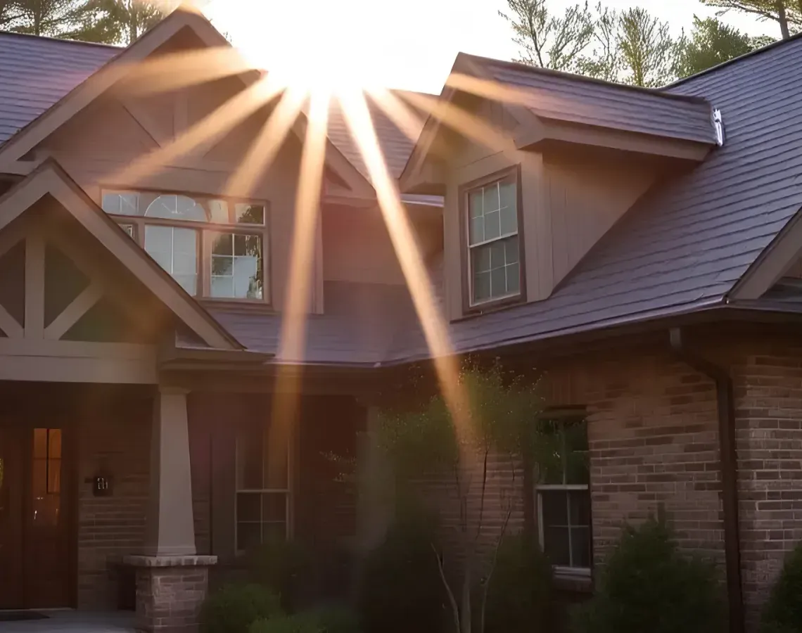 Sunlight streaming over a brick house with a steep roof and front porch at sunset
