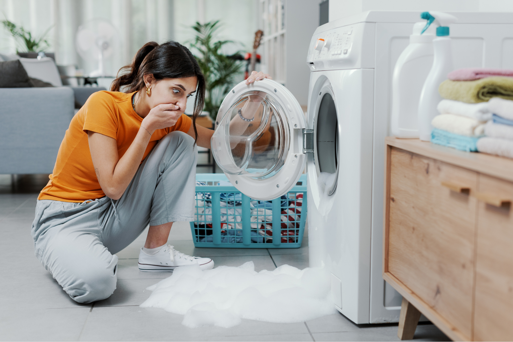 Woman kneels beside a washing machine with the door open, soapy water on the floor. She appears disgusted.