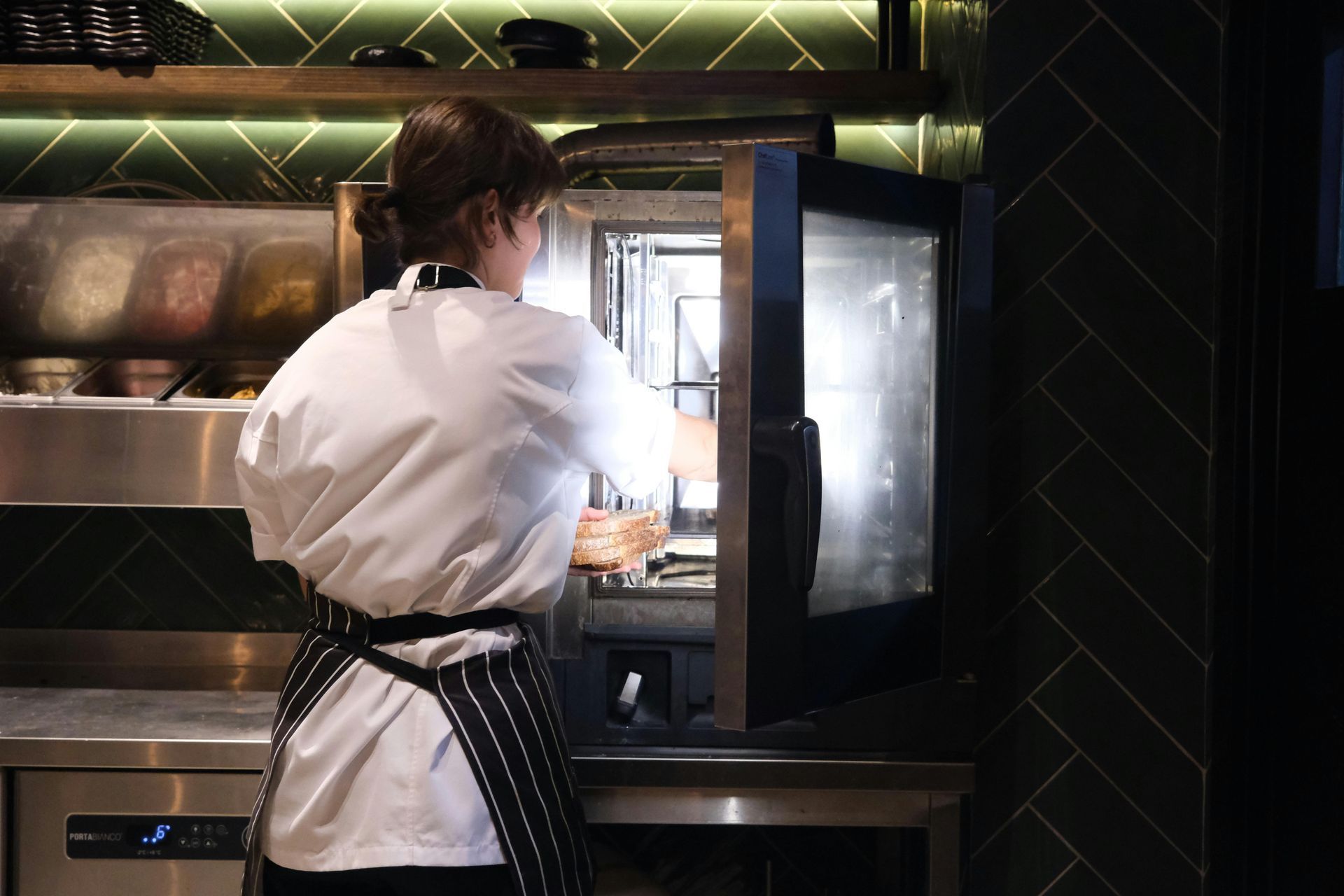 Chef in a white coat and apron opening a commercial oven in a restaurant kitchen.