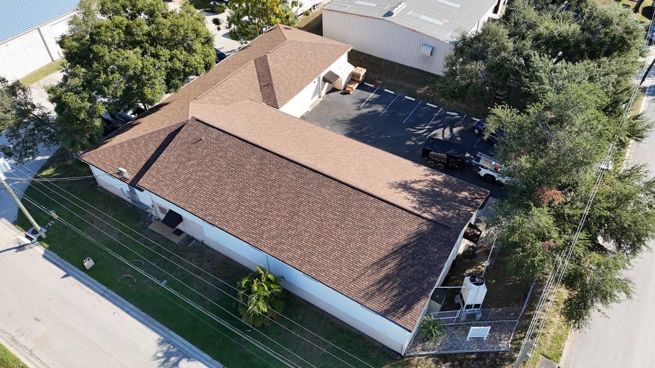 Aerial view of a brown-roofed, white building in a tree-lined lot, with a driveway and green lawn.