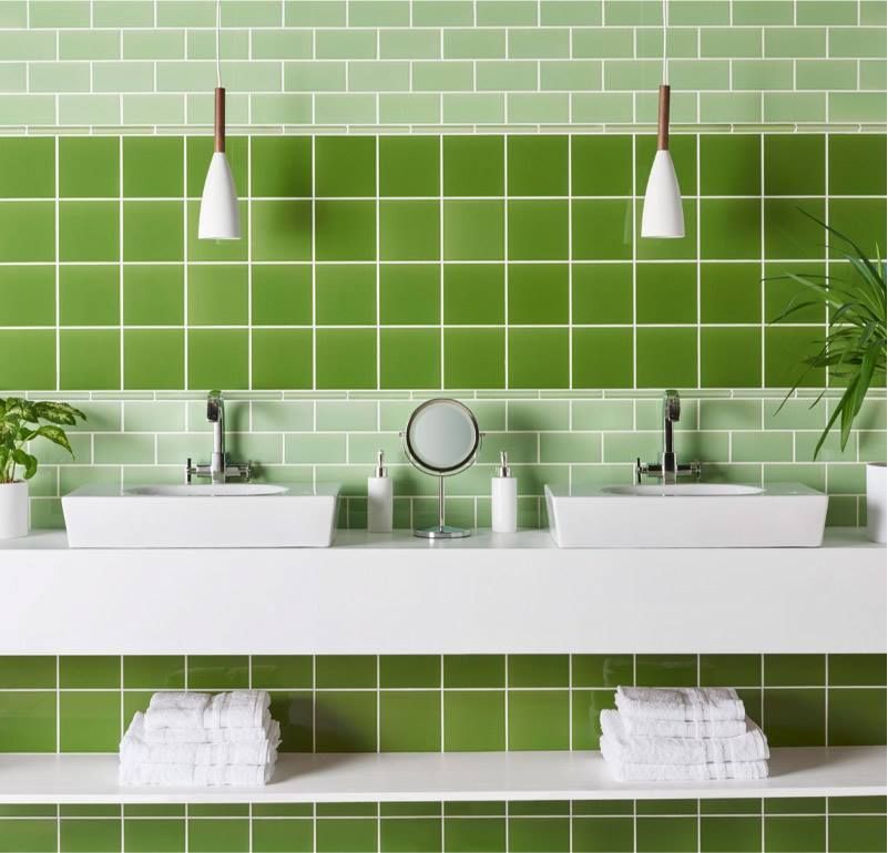 Bagno piastrellato di verde con lavandini bianchi, asciugamani e piante decorative. Due lampade a sospensione sono appese sopra i lavandini.
