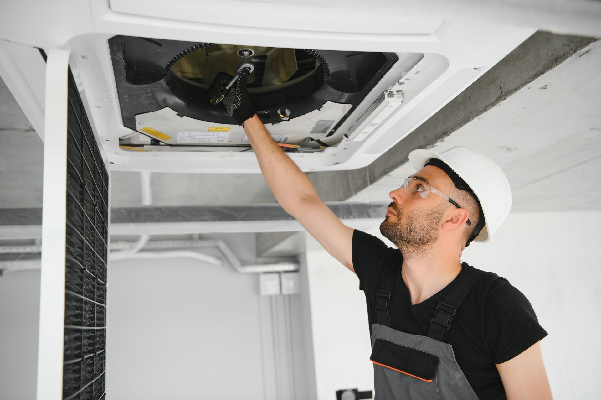 HVAC technician in a white hard hat inspects an air vent, reaching up, indoors.