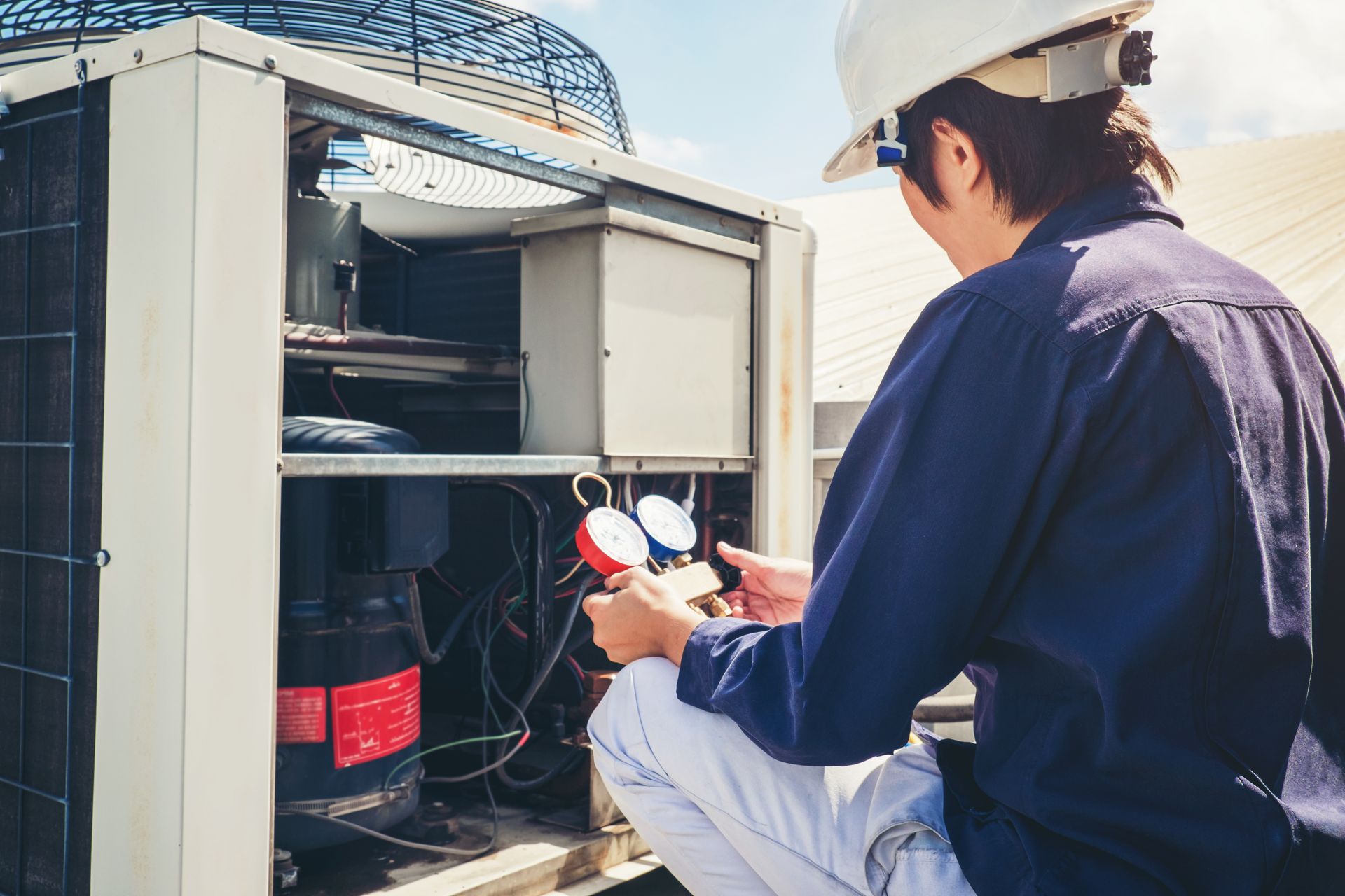 HVAC technician in a white helmet and blue jumpsuit checks gauges on an air conditioning unit on a rooftop.