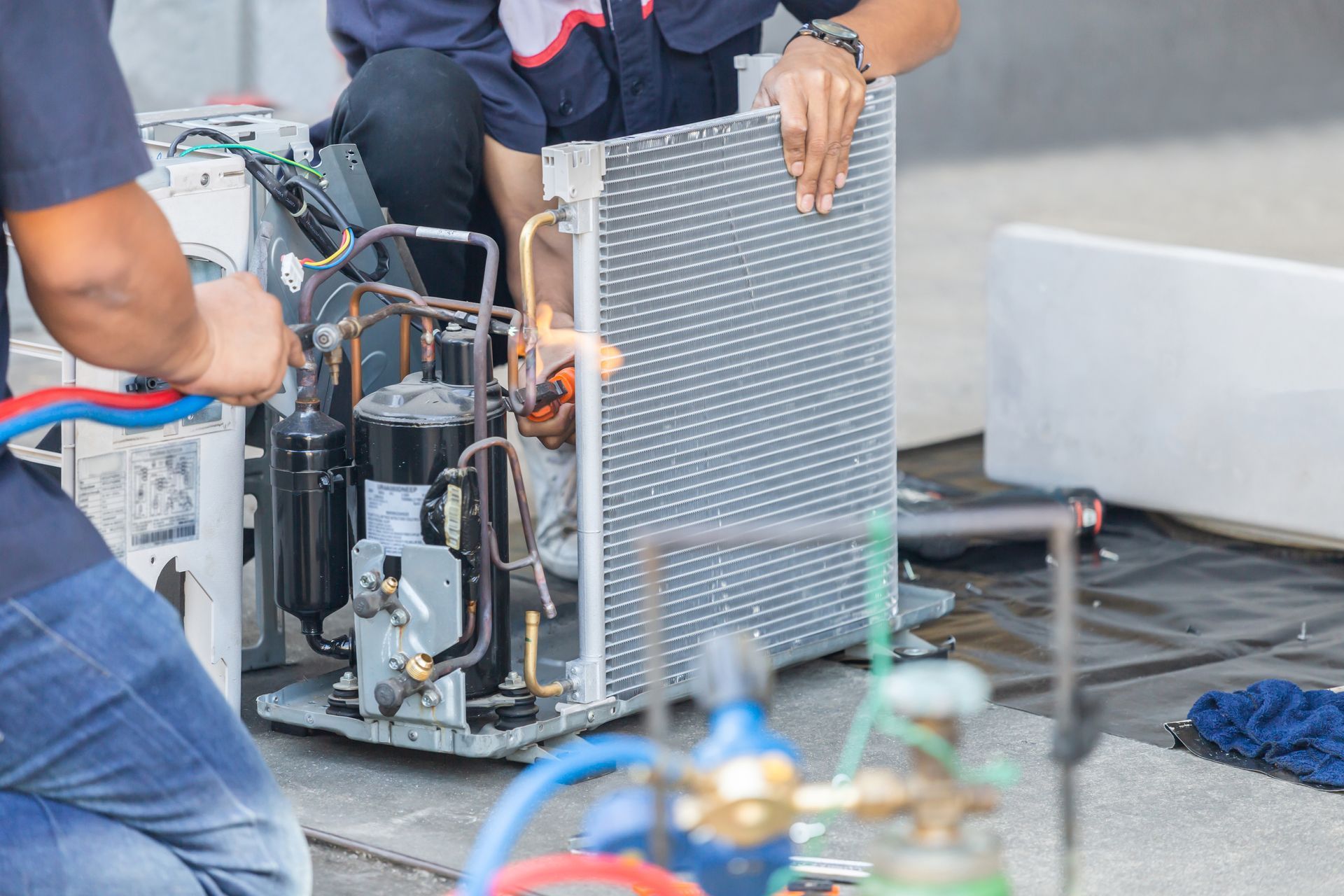 HVAC technicians repairing air conditioning unit outdoors. One holds radiator, other works on pipes.