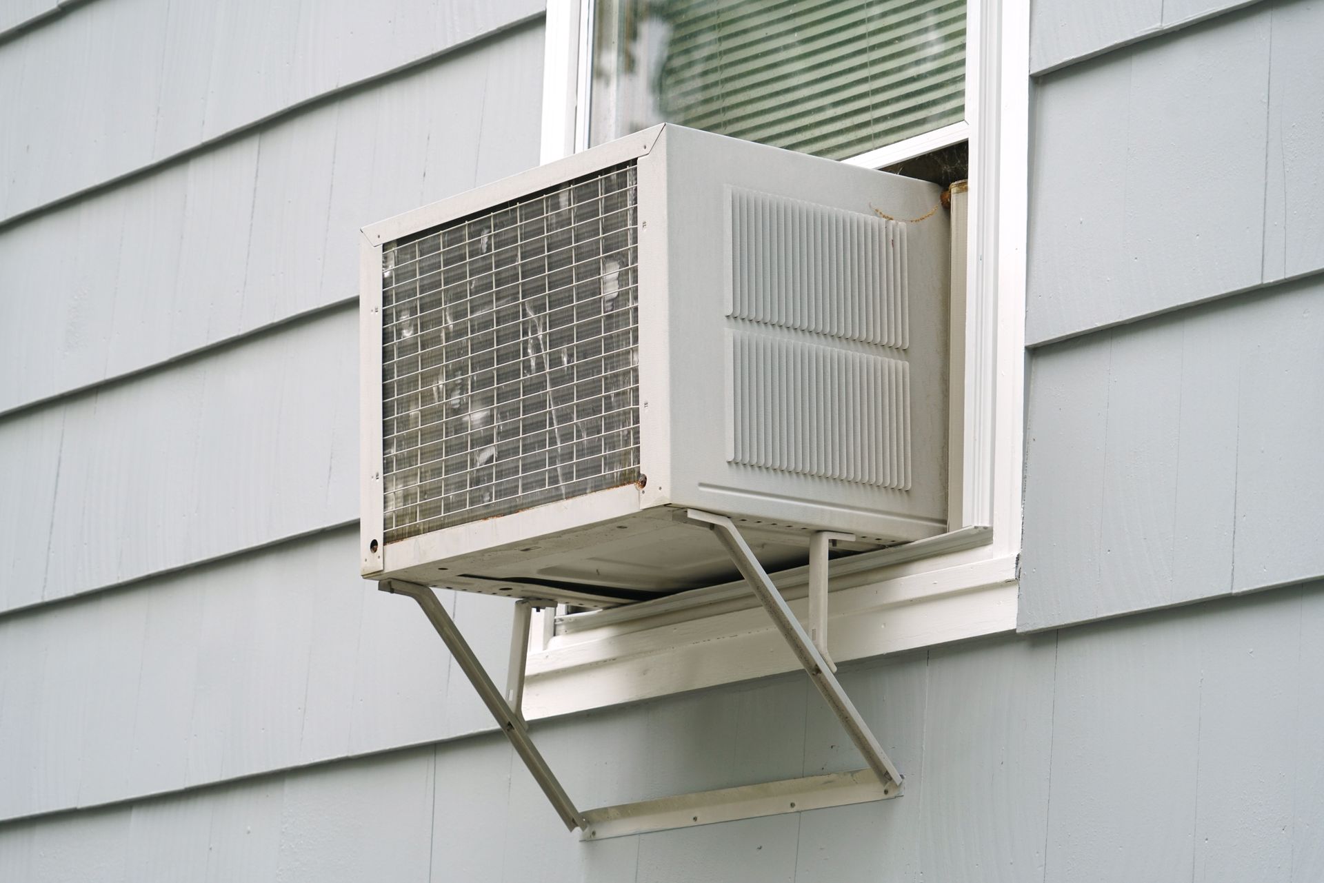 Window air conditioner unit installed in an open window, exterior view. White siding and support brackets are visible.