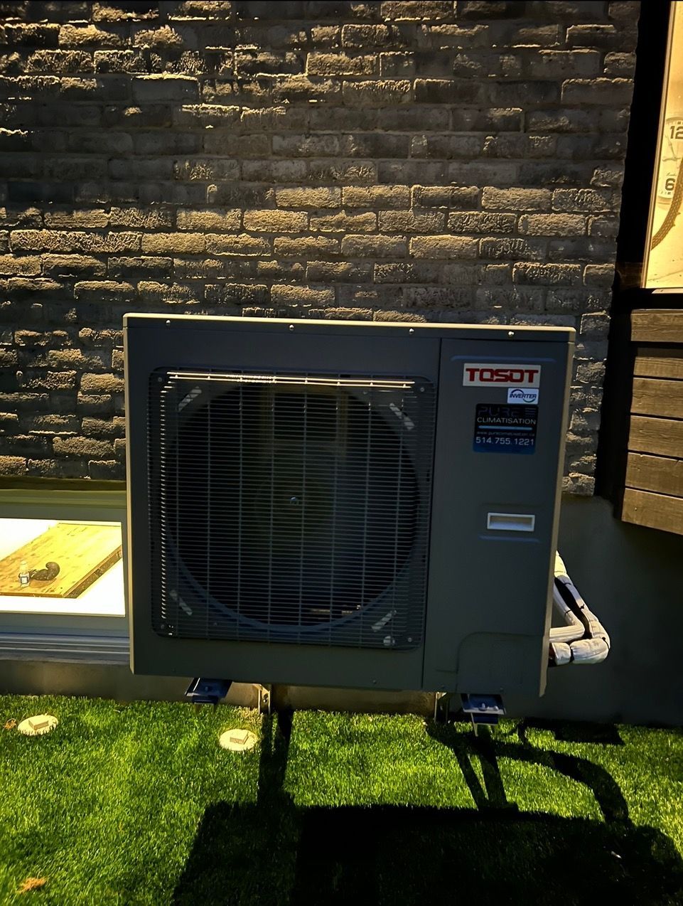 Un climatiseur est posé sur l'herbe devant un mur de briques.