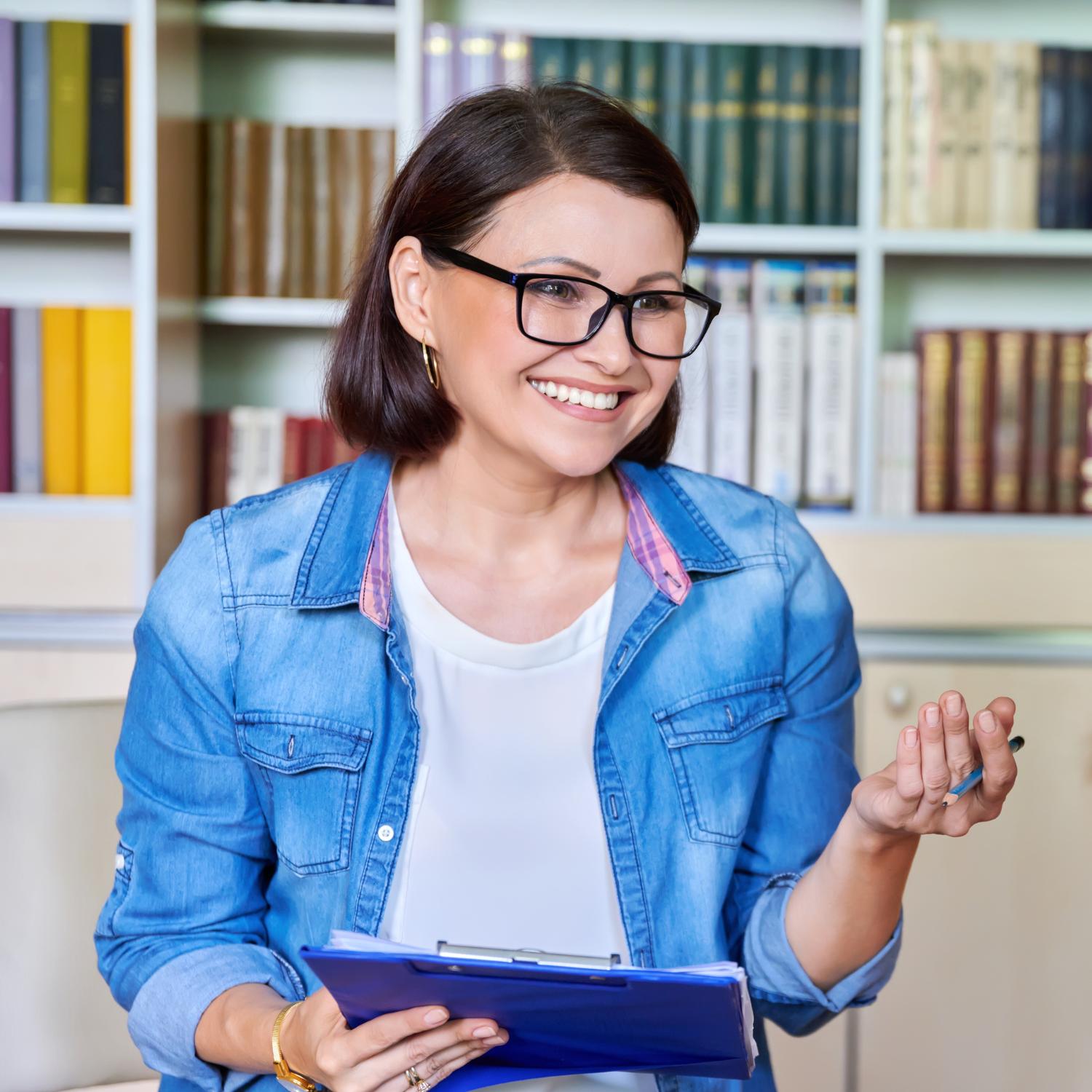 A woman wearing glasses and a denim jacket is holding a clipboard and smiling.