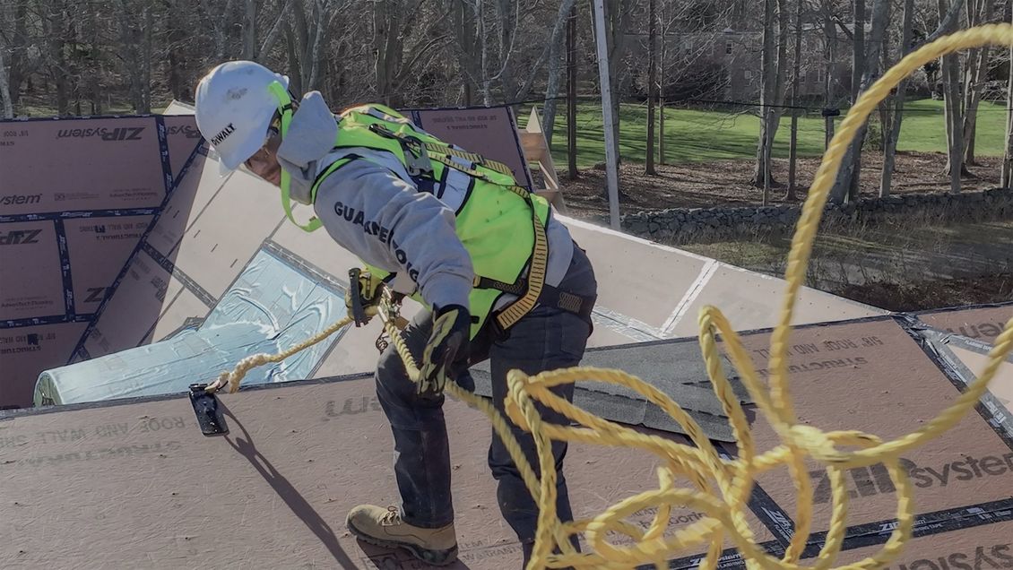 Roofer on a pitched roof wearing safety gear, including a helmet and harness, working with a rope.