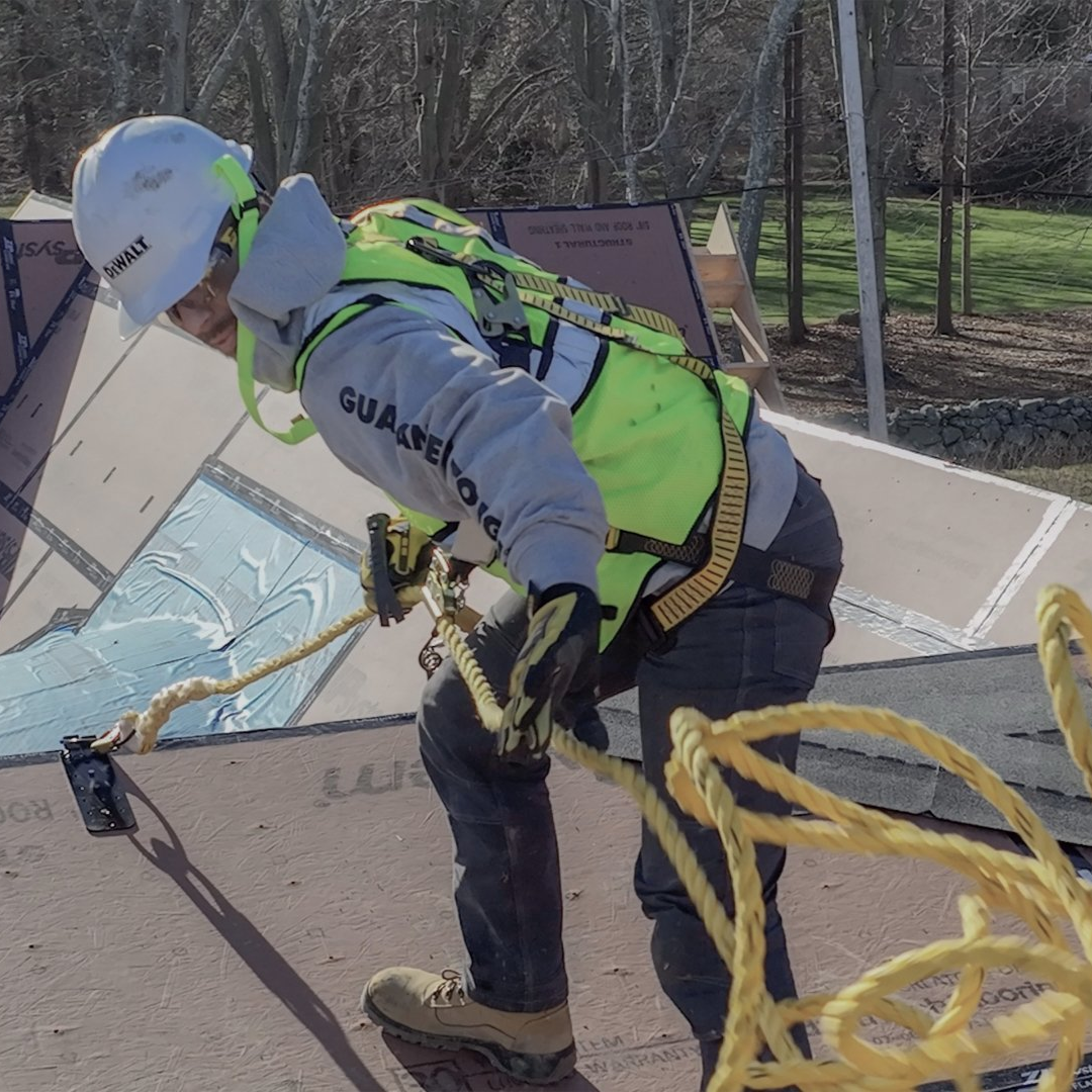 Roofer on a pitched roof wearing safety gear, including a helmet and harness, working with a rope.