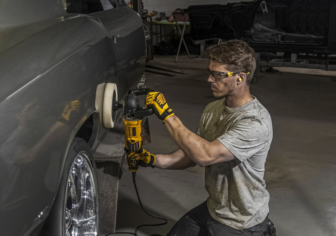 Man using a polisher on a car's fender, wearing safety glasses and gloves in a workshop setting.