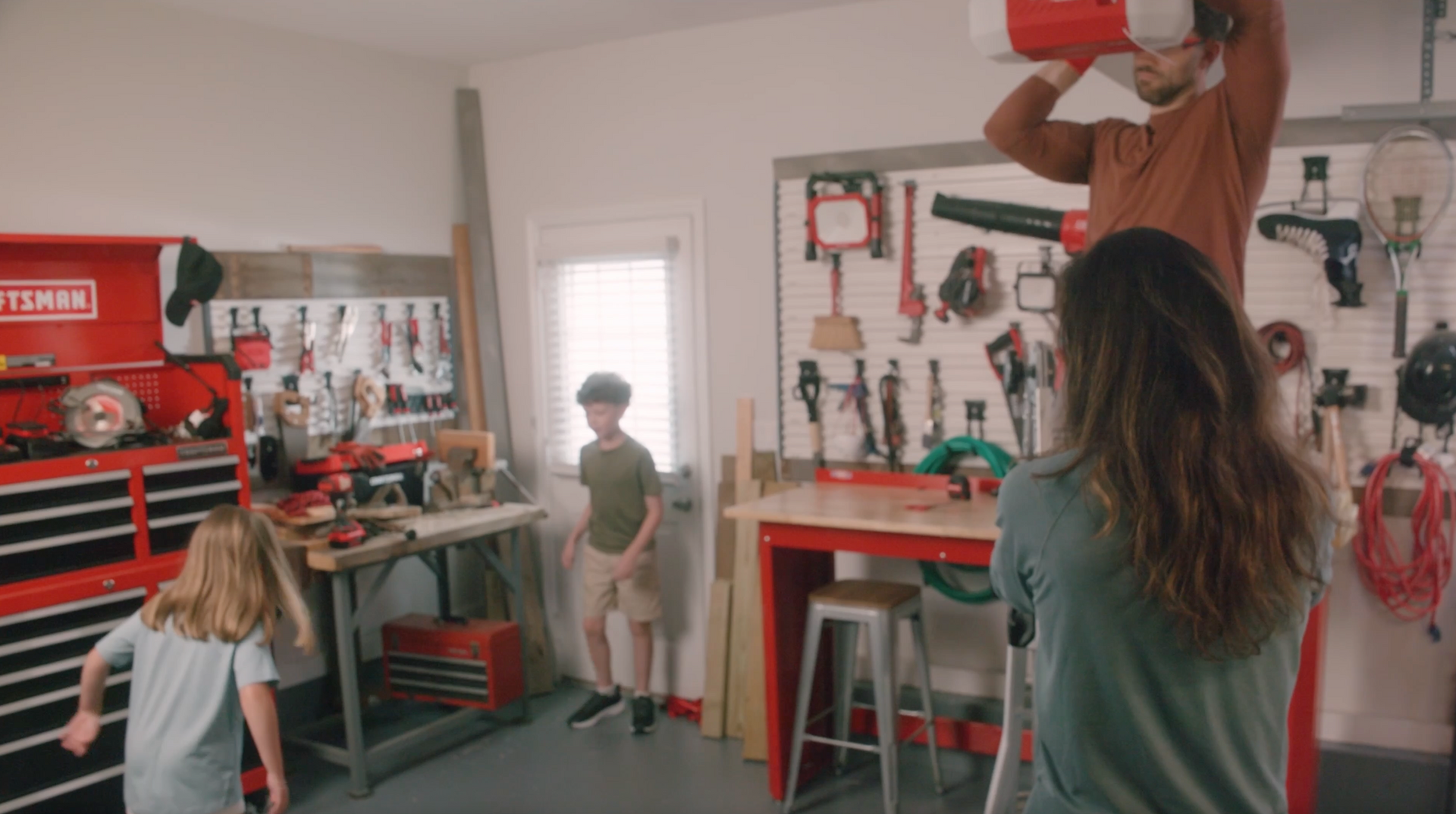 Family in a garage. Dad on a ladder, lifting. Boy and girl watching. Tools on wall and workbench.