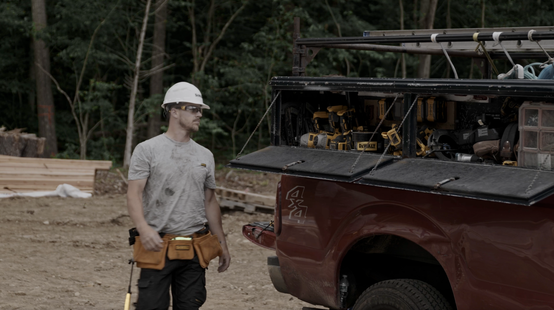 Construction worker with tool belt and hard hat stands by red truck with open tool storage.