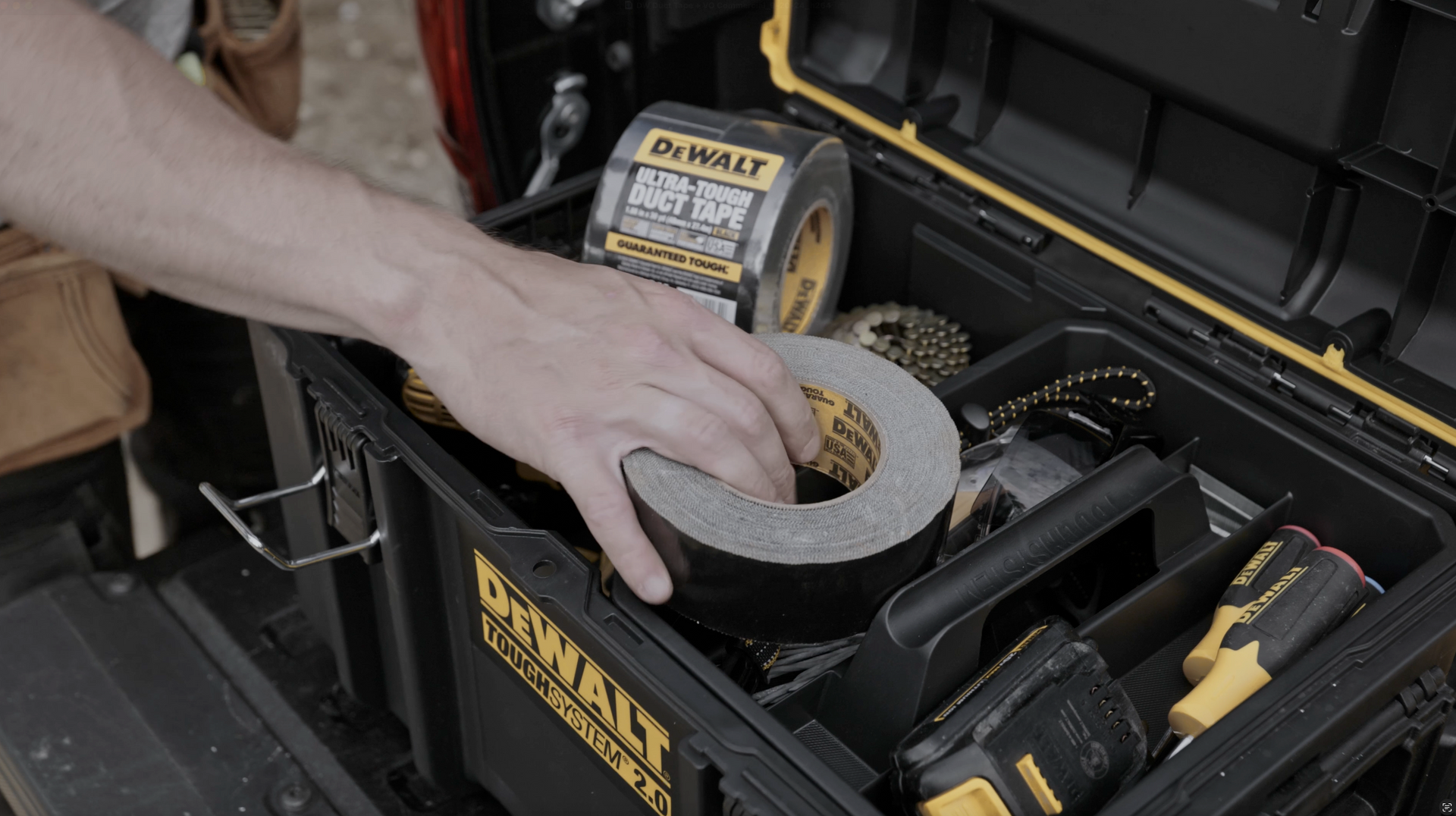 Hand reaching into a black and yellow tool box for a roll of gray duct tape.