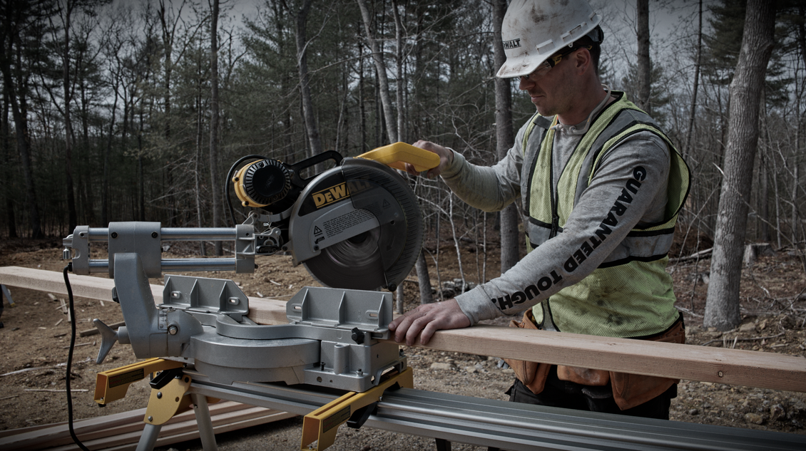Construction worker using a miter saw outdoors, cutting wood. He wears a hard hat and safety vest.