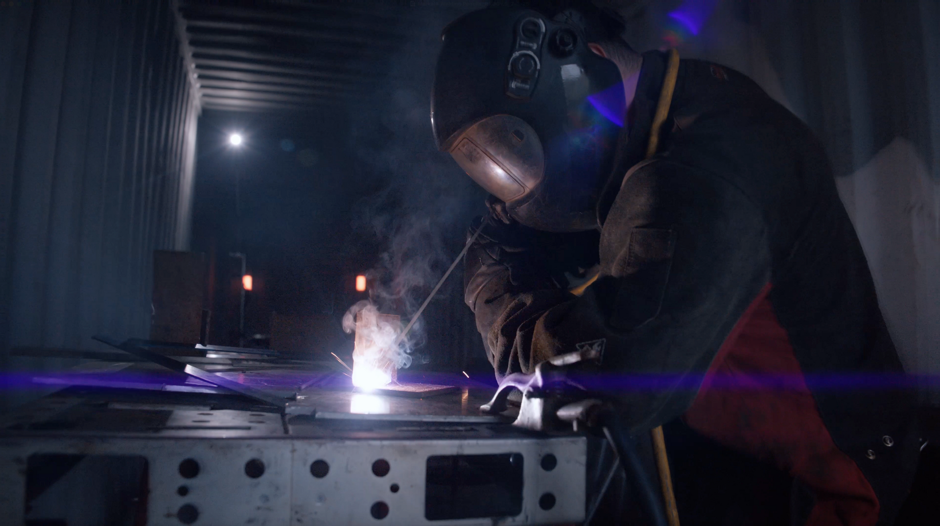 Welder wearing protective gear, welding metal on a workbench inside a dark workshop.