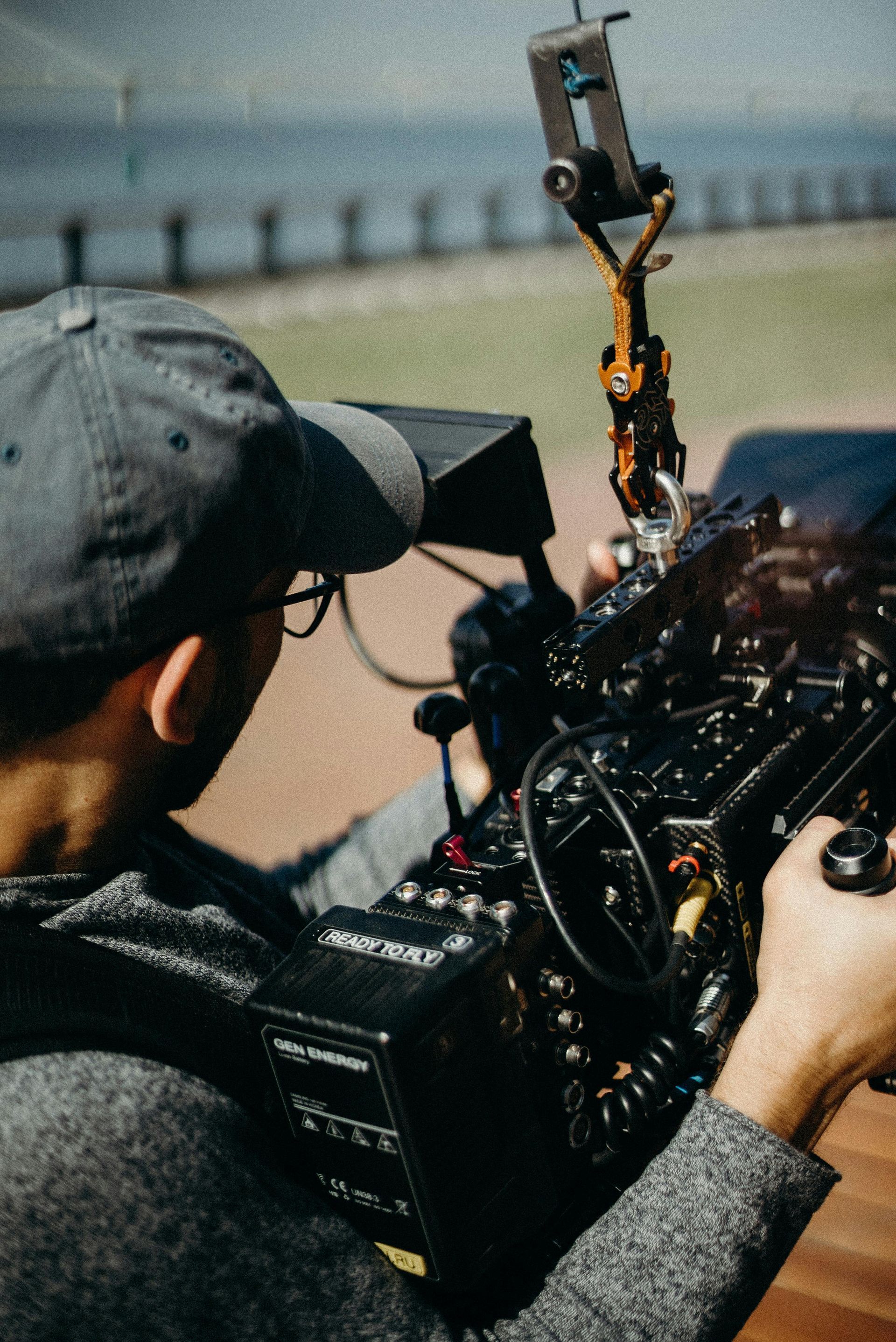A person in a gray cap operates a large professional video camera with a blurred outdoor backdrop.