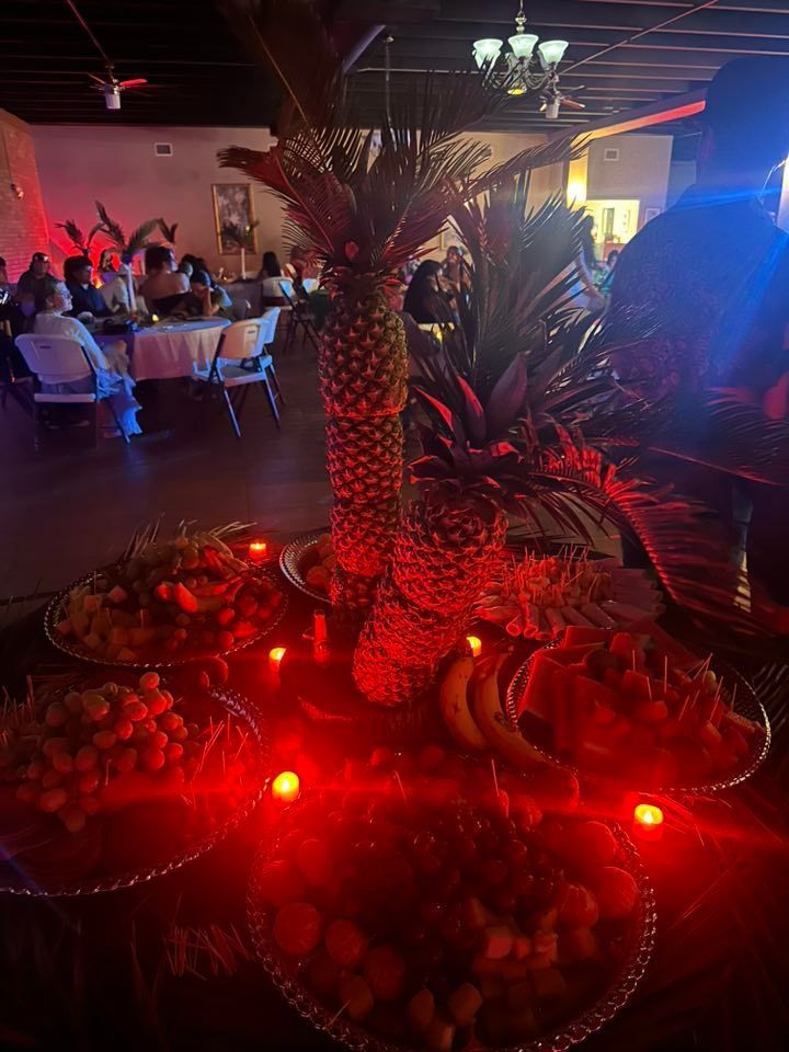 Fruit and decorative palm arrangement on table with red lighting. Guests in background.