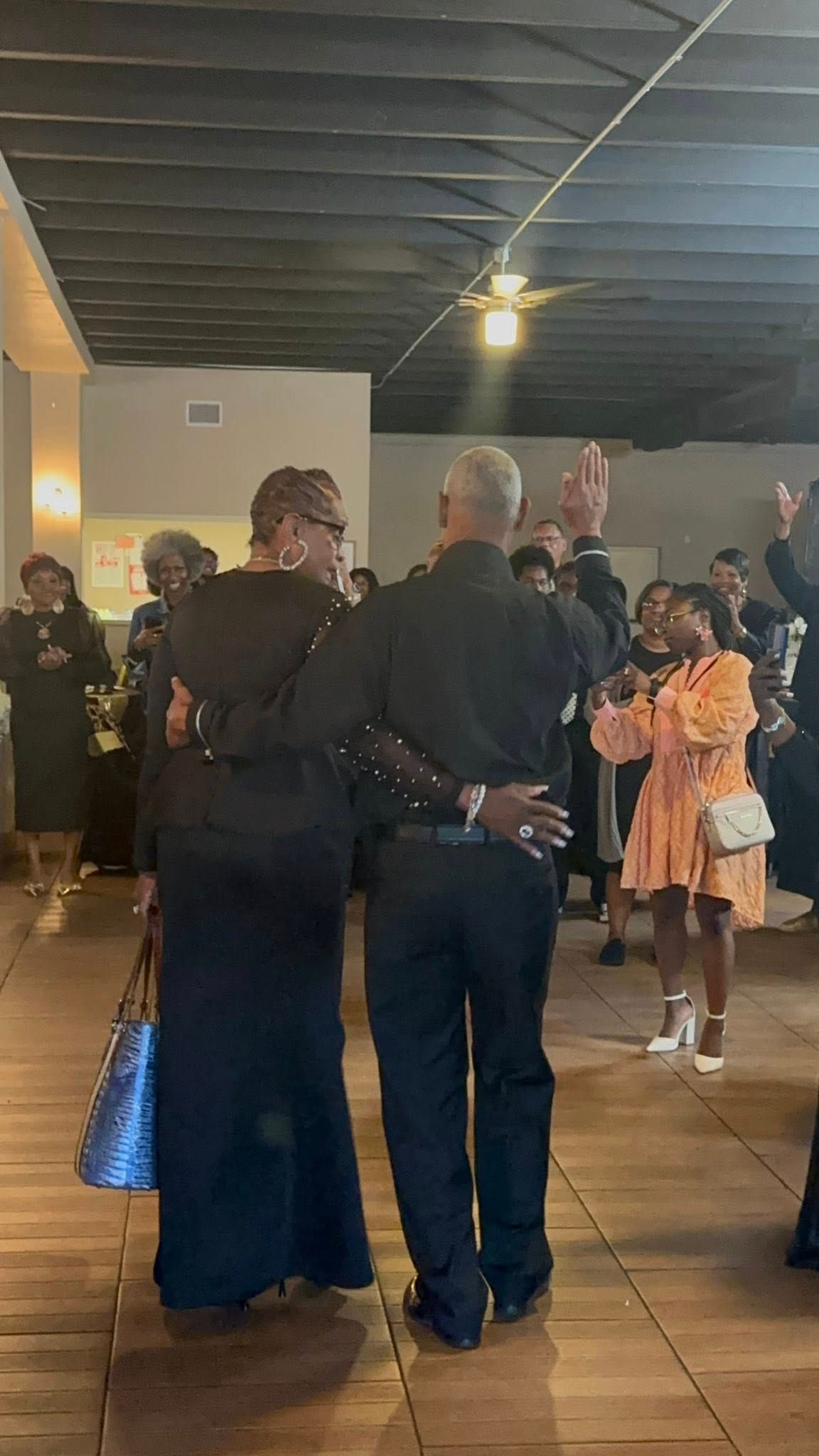 Couple dancing, embraced. Others watch, some with hands raised in a room.