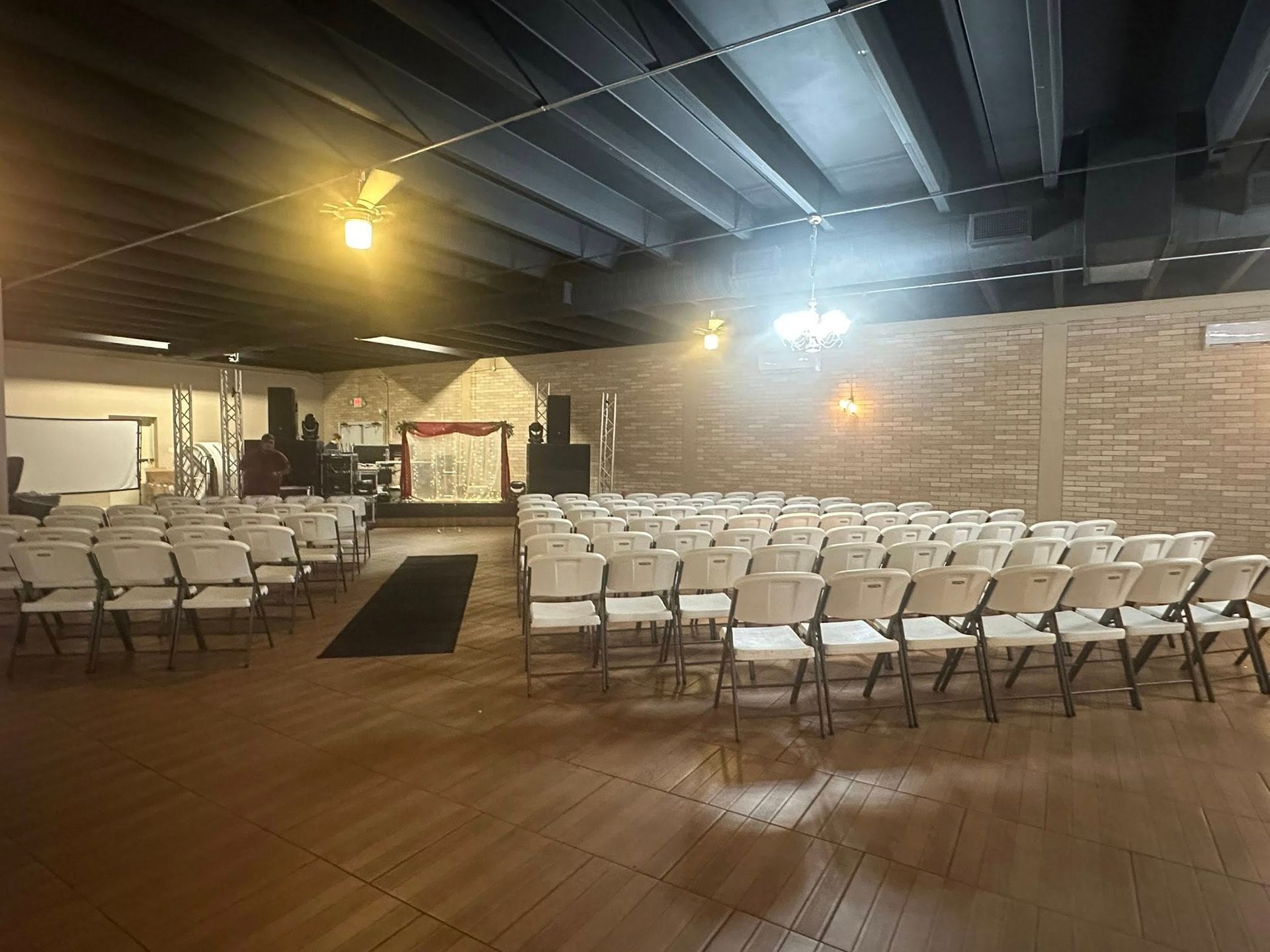 An indoor event space with white folding chairs arranged for seating, facing a stage area. Brick walls and dark ceiling.