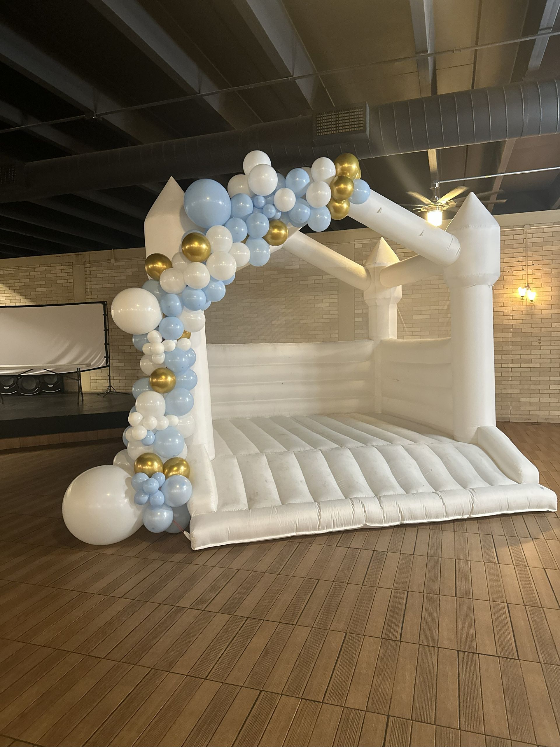 White bounce house with blue and gold balloon arch, indoors.