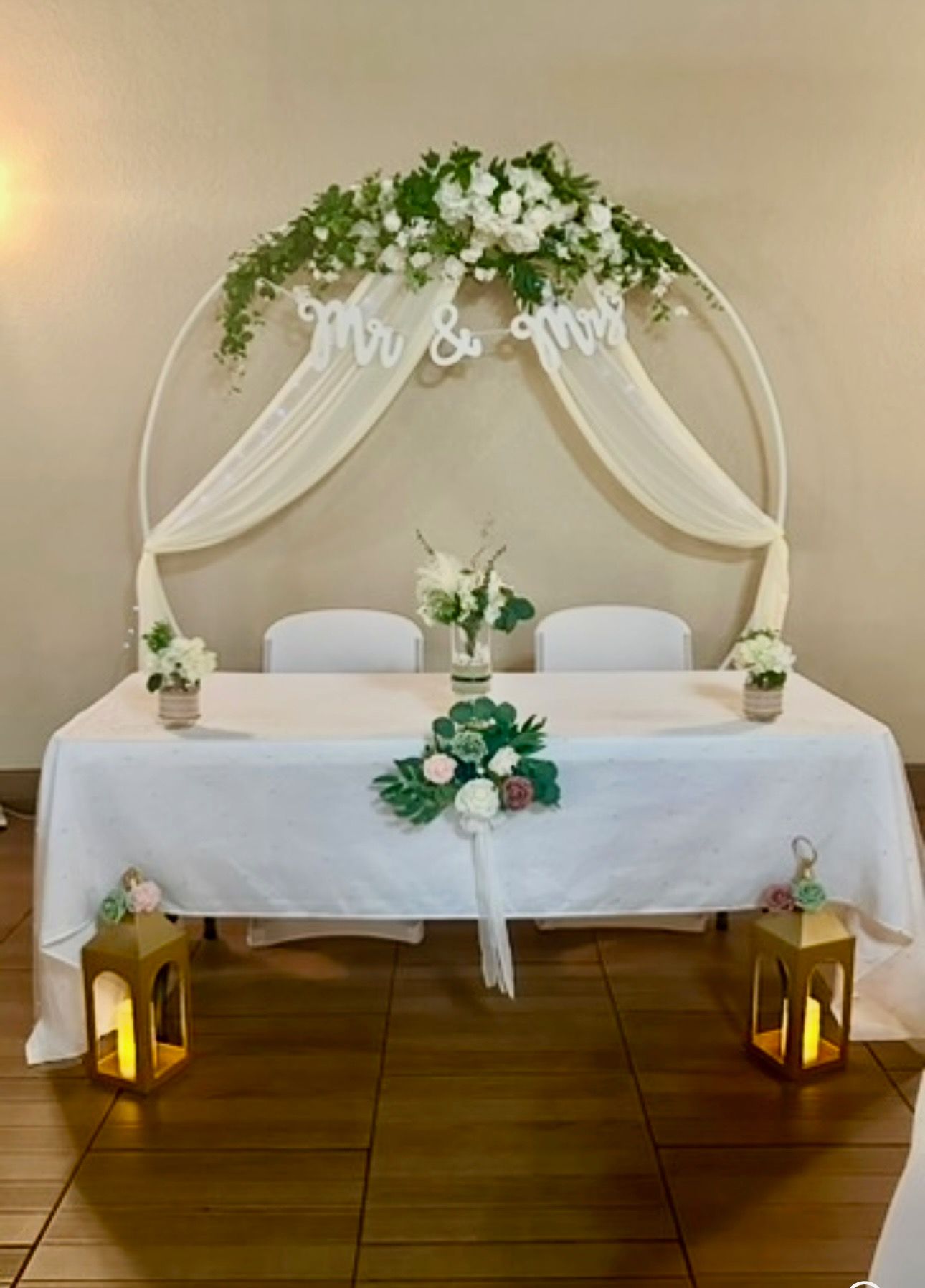 Wedding sweetheart table with white draping, floral arch, and lanterns.