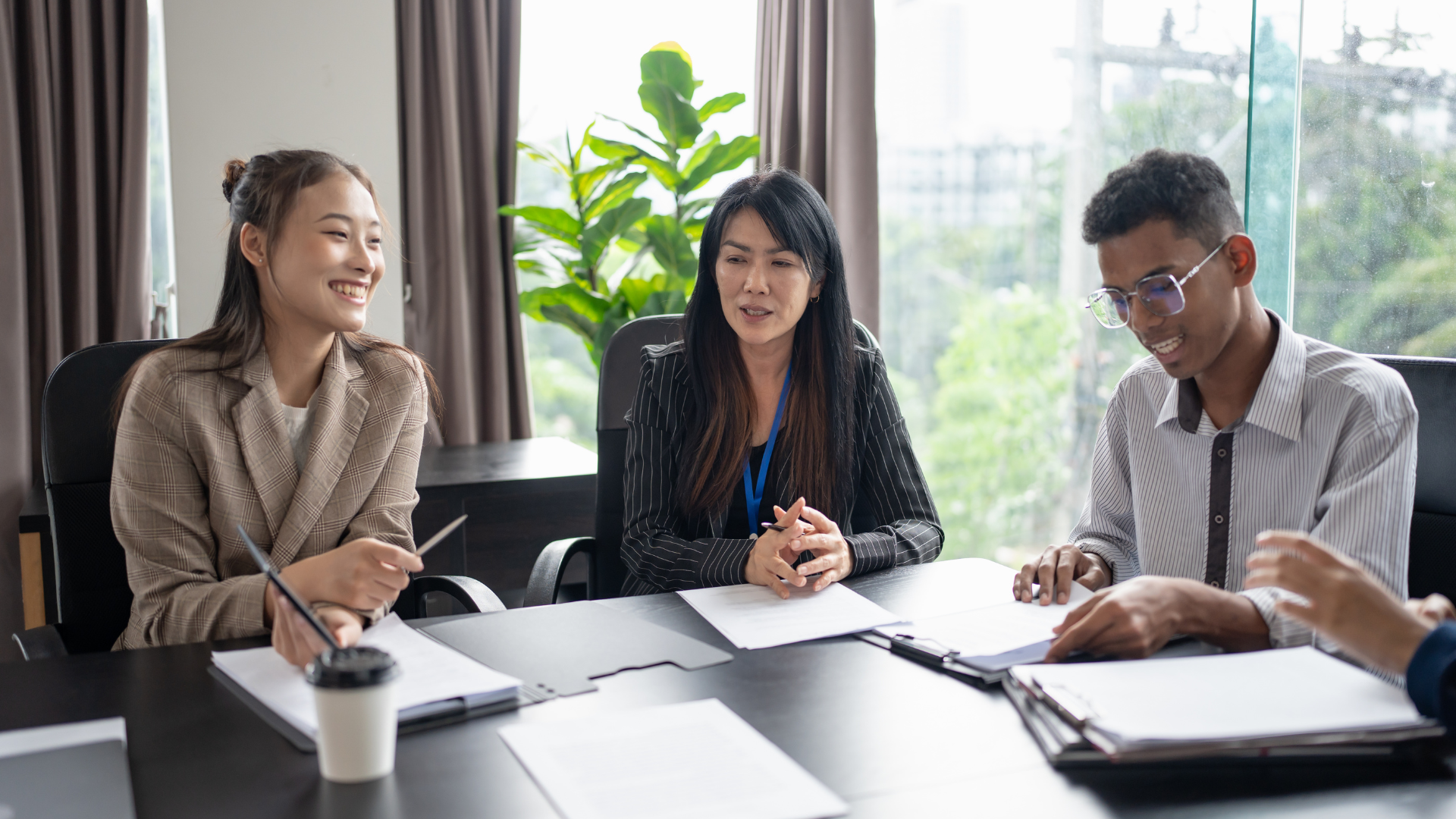 Three colleagues in a meeting room discussing documents at a table, with laptops and notebooks.