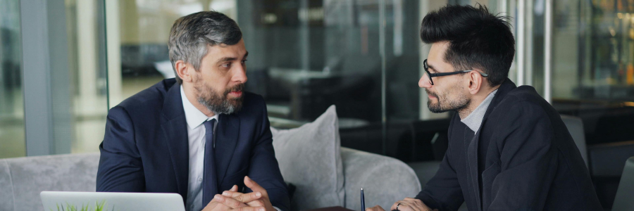 Two men in suits talking, seated in a modern, well-lit area with reflective glass.