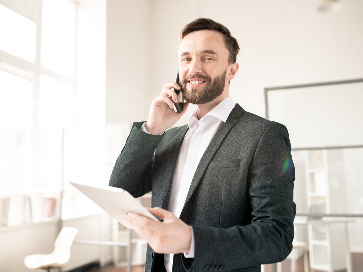 Man in suit on phone, holding tablet, smiling in office setting.