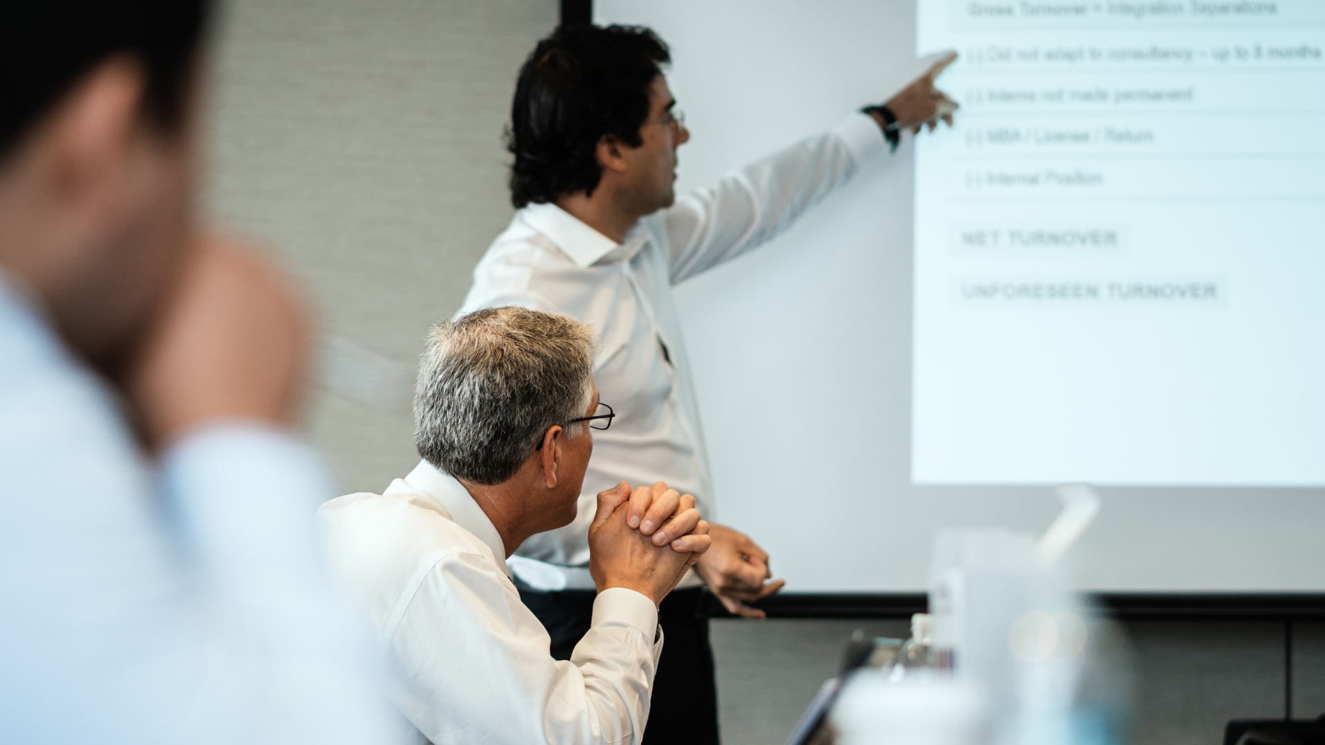 Man in suit shaking hands with another man, woman smiles nearby in a car dealership.