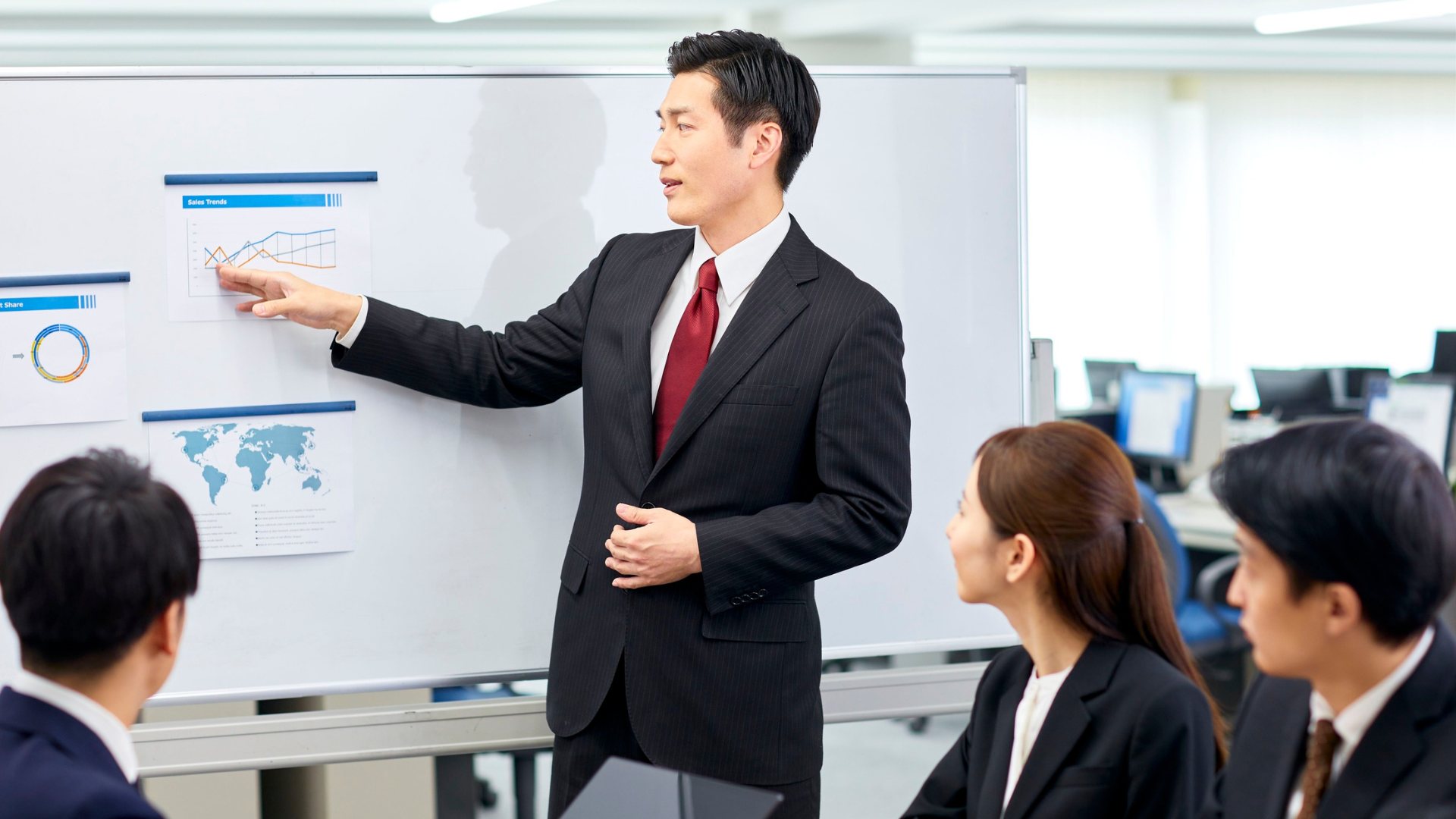 Man in suit presenting graphs on whiteboard to colleagues in an office.