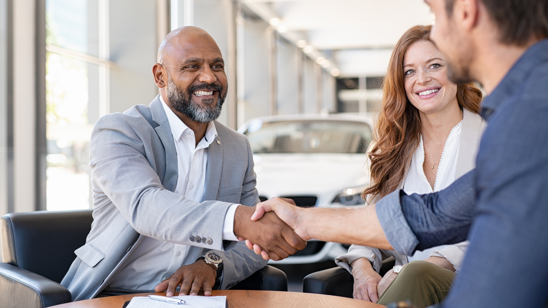 Man in suit shaking hands with another man in a car dealership, a smiling woman sits nearby.