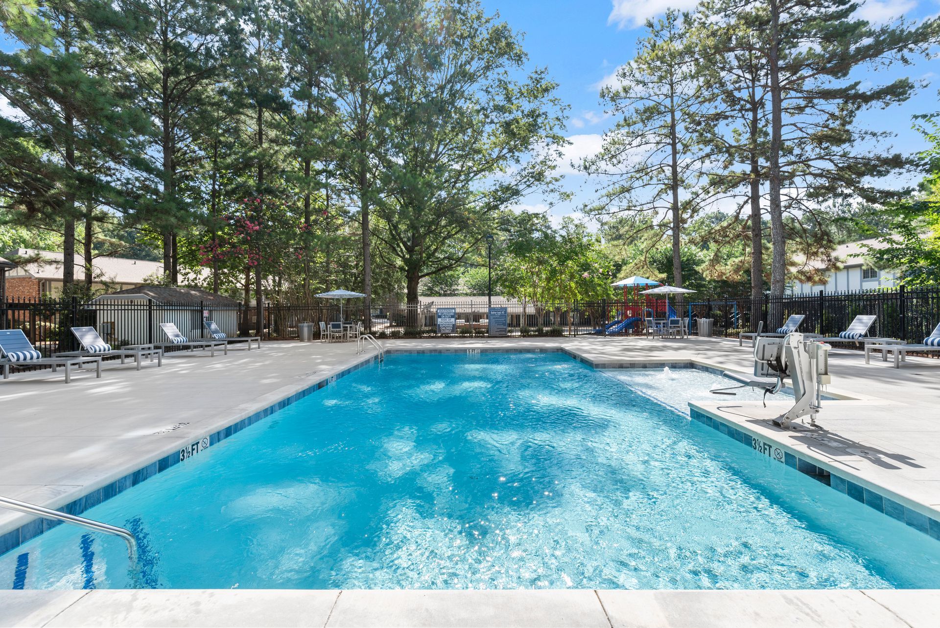 Pool with blue water and surrounding deck, lounge chairs, trees, and buildings under a blue sky.
