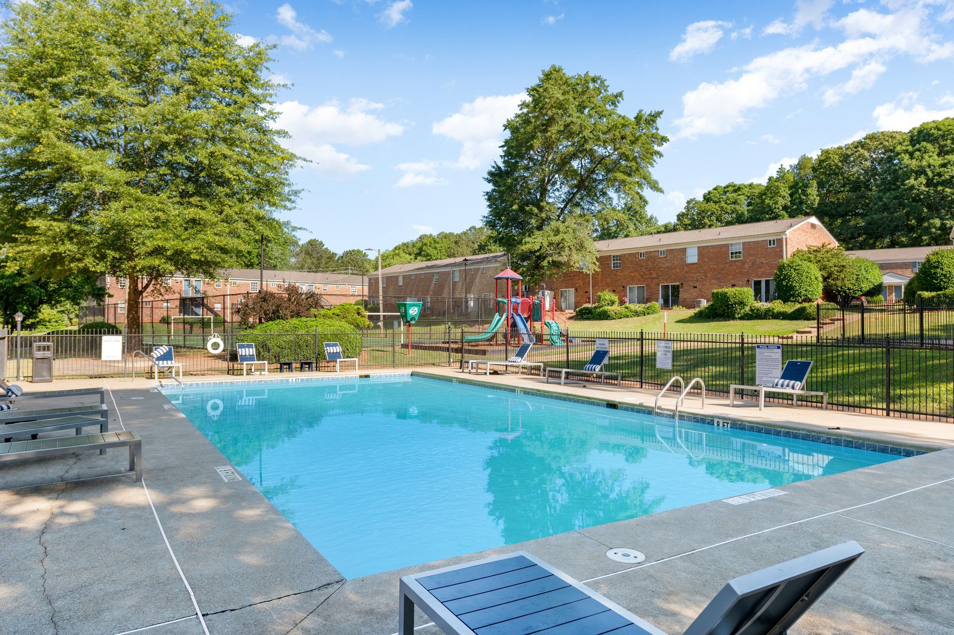 Swimming pool with lounge chairs, playground in the background, apartment buildings, and trees under a blue sky at The Arbors at Smyrna, offers apartments near The Battery in Smyrna, GA.
