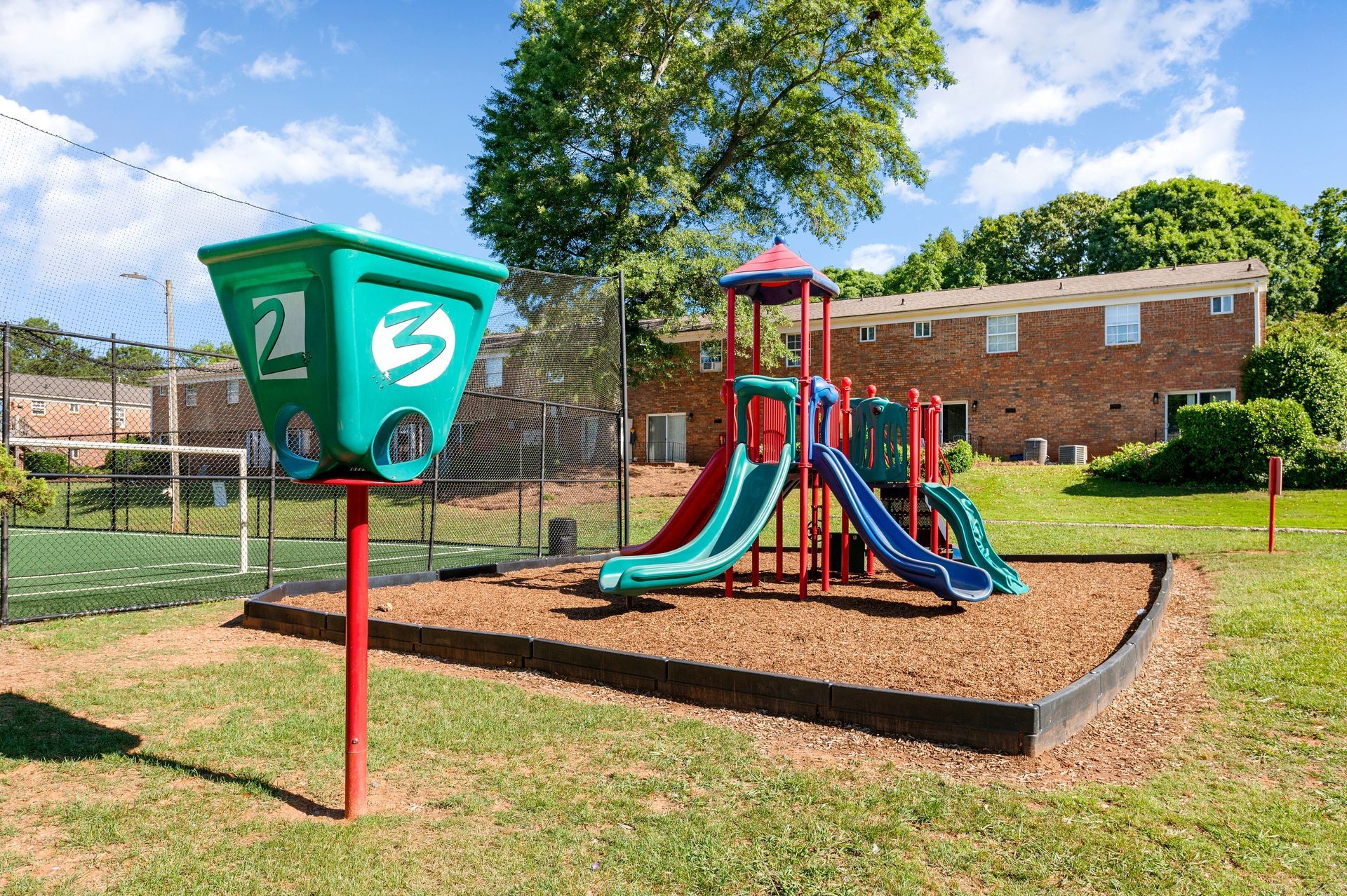 Playground with green basket for shooting, slides, and brick apartments in the background at The Arbors at Smyrna, offers townhomes in Smyrna, GA.