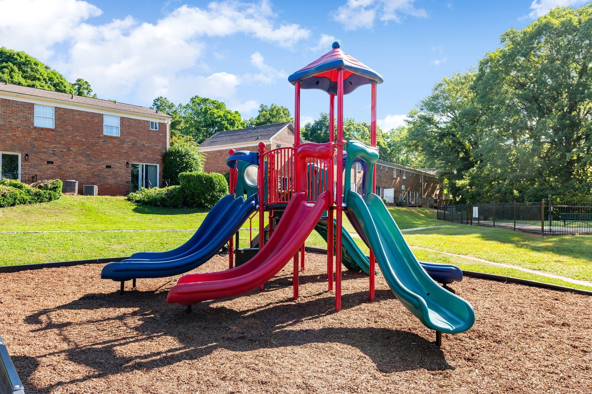 Playground with blue, red, and green slides on wood chips at The Arbors at Smyrna, offers townhomes in Smyrna, GA. Brick apartment  and townhome buildings in the background.
