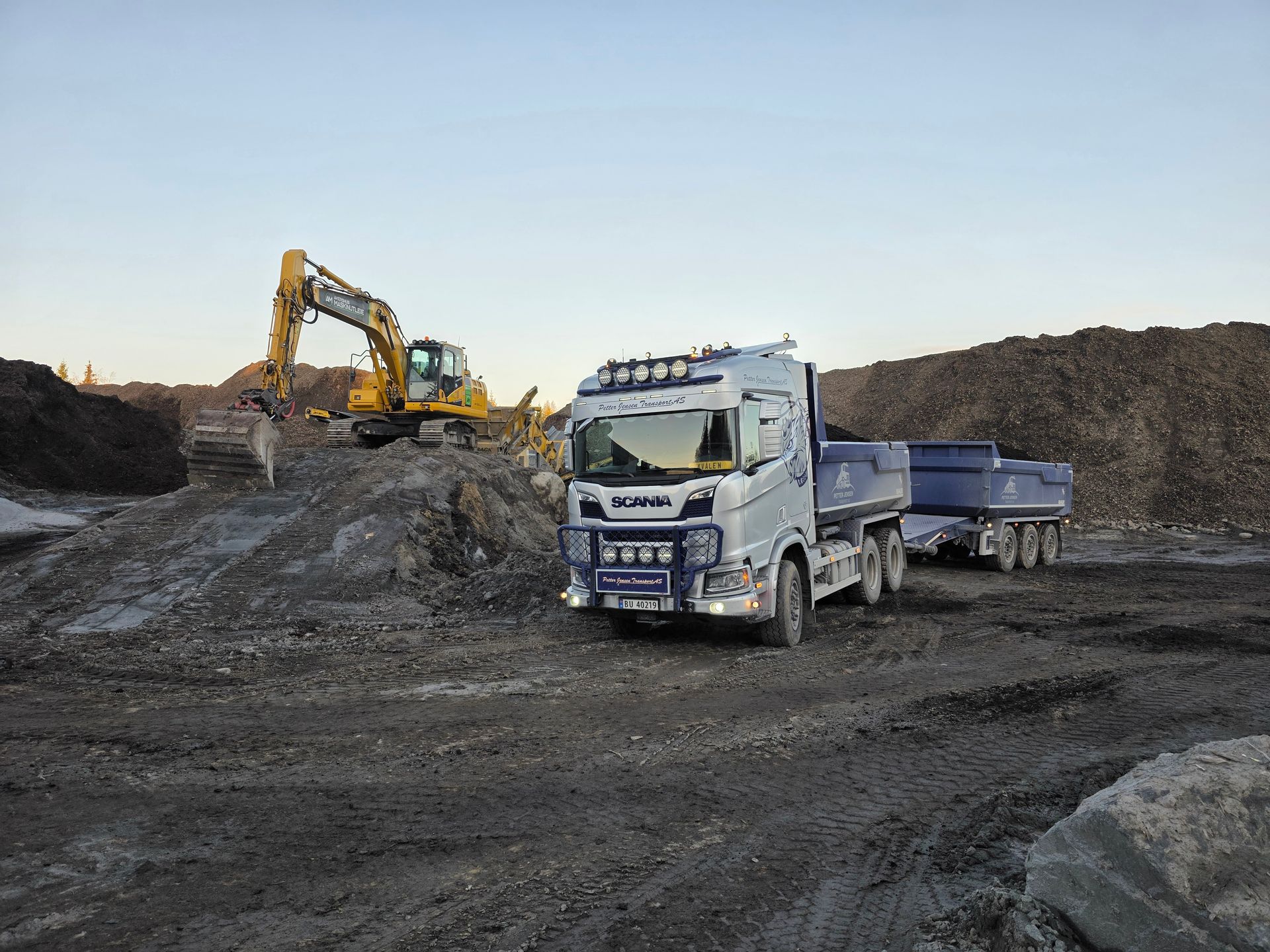 En blågrønn gravemaskin laster en blå dumper med jord. Sett mot en lys blå himmel og spredte grønne trær.